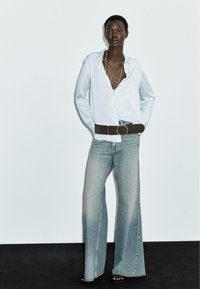 Camicia azzurra chiara, vestibilità morbida e sbottonata in alto. Jeans in denim chiaro a vita alta e a gamba larga. Cintura marrone con fibbia circolare.