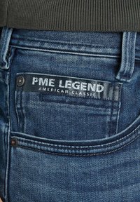 Jeans de denim azul con un acabado texturizado. Presentan una etiqueta negra con el texto "PME LEGEND AMERICAN CLASSIC", y detalles de botones metálicos.