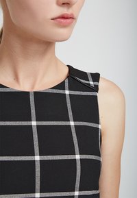 Top sans manches à carreaux noir et blanc avec un col structuré et une texture douce. Couture visible et détails de lignes.