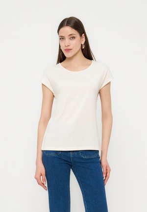 Mujer con cabello largo y castaño, que lleva pendientes de aro dorados, una camiseta blanca de manga corta y vaqueros azules, de pie frente a un fondo liso.