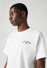 Vit bomulls t-shirt med korta ärmar, som har en böjd svart textgrafik "LA VILLE LUMIÈRE" och en liten röd acccent på bröstet.