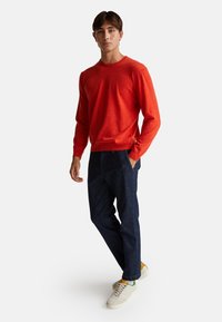 Mężczyzna w czerwonym swetrze, ciemnoniebieskich dżinsach i białych sneakersach z musztardowymi akcentami, na jednolitym jasnym tle.