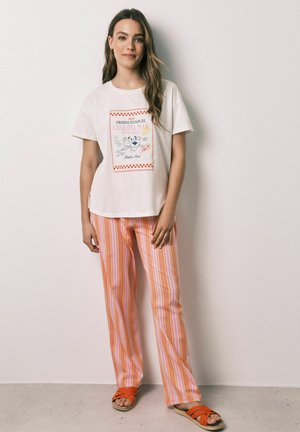 Vrouw met lang haar die een wit bedrukt T-shirt, oranje en roze gestreepte broek en oranje sandalen draagt, staand tegen een effen witte muur.
