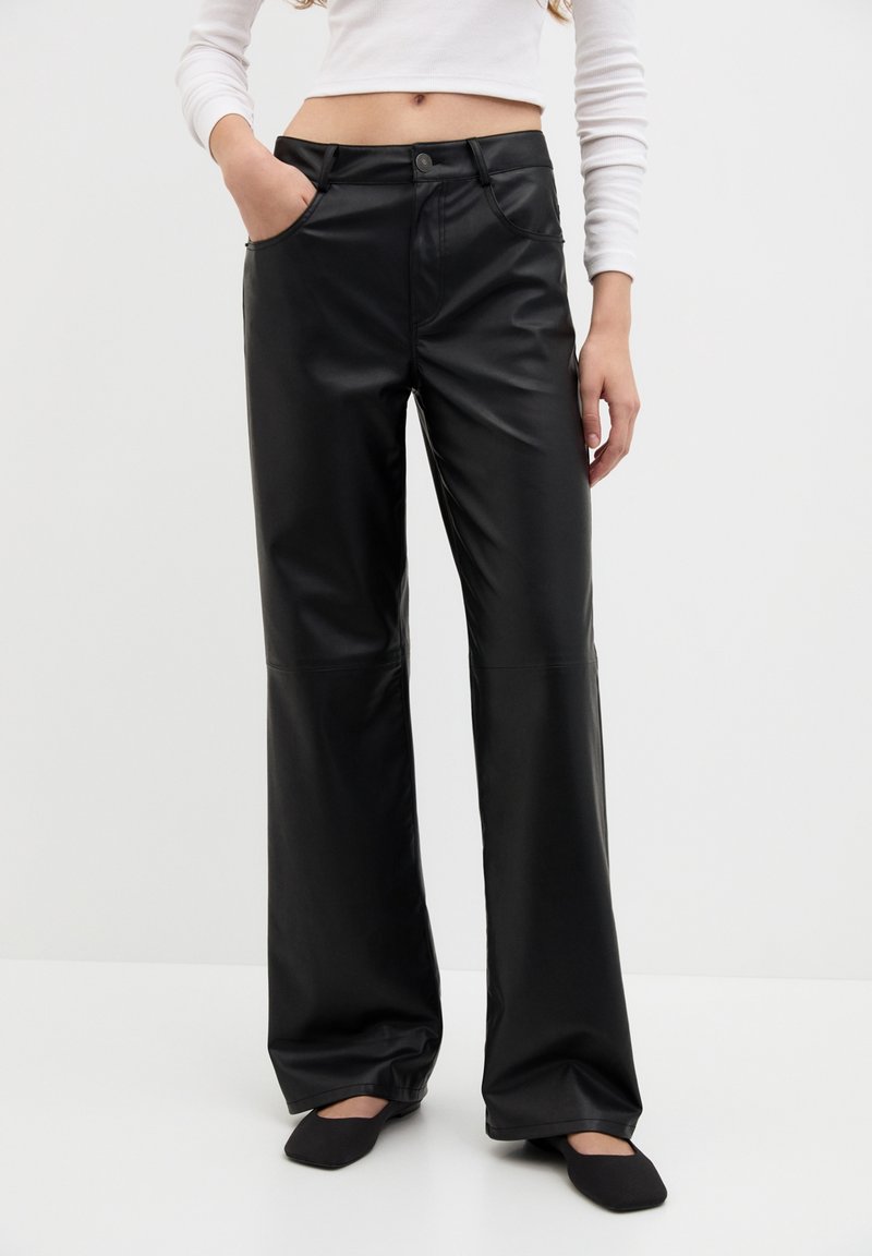 Femme portant un pantalon large en faux cuir noir, un haut court à manches longues blanc et des chaussures noires à bout carré, se tenant avec une main dans sa poche.