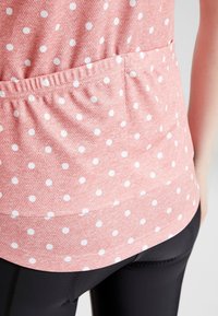 Różowa bluzka w białe kropki, wykonana z miękkiego materiału, z elastycznym paskiem w talii i zaokrąglonym dołem. W zestawie z czarnymi legginsami.