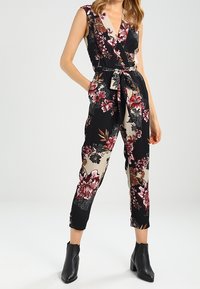 Svart blommig jumpsuit med djup v-ringning, insydd midja och korta ben. Har stora rosa och krämfärgade blommor på en kontrasterande bakgrund.