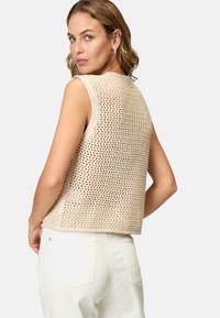 Beige hæklede ærmeløs top med et åbent vævemønster. Kasseformet design med rund halsudskæring. Kombineret med hvide jeans.