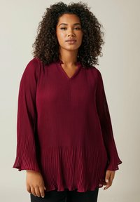 Blouse plissée bordeaux avec un col en V, des manches longues et un ourlet évasé. Présente une texture verticale et un tissu léger.