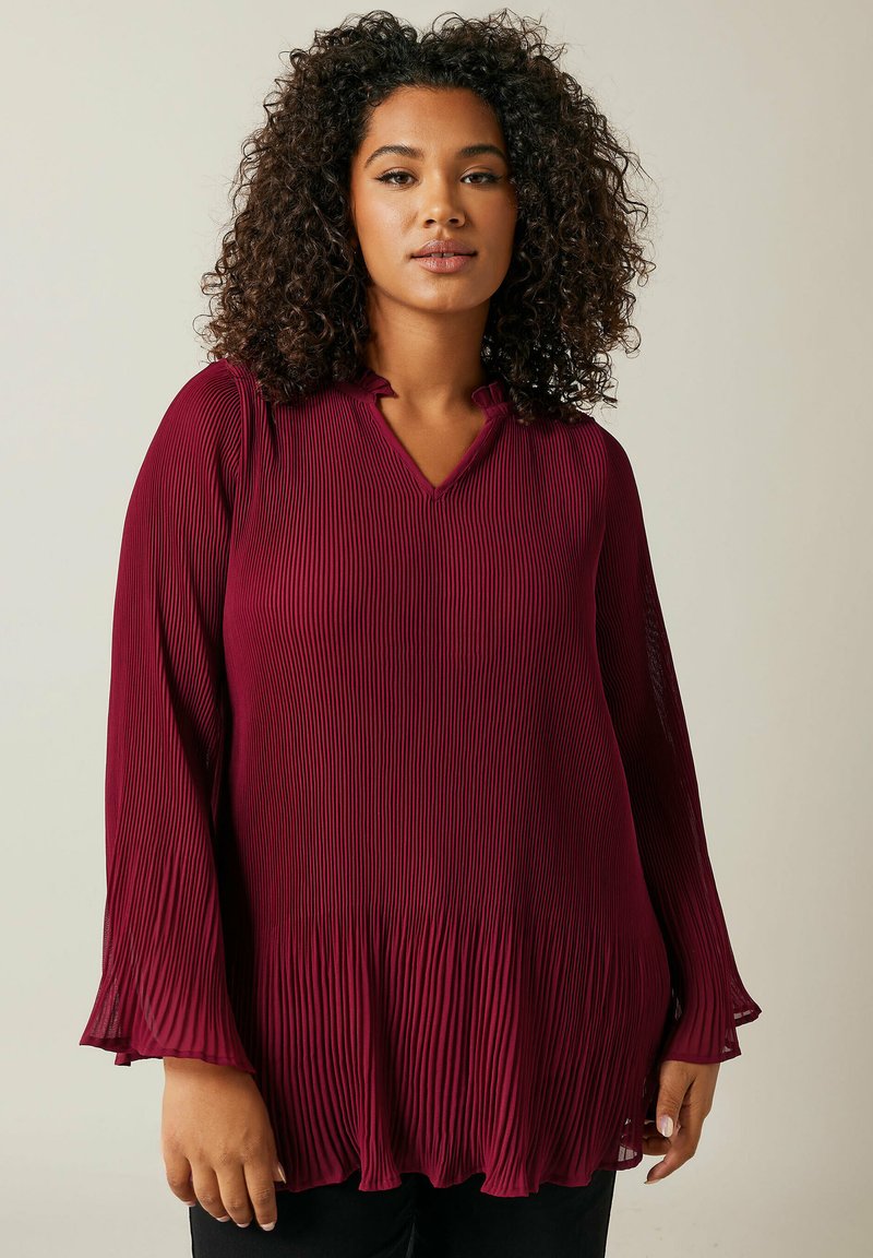 Blouse plissée bordeaux avec un col en V, des manches longues et un ourlet évasé. Présente une texture verticale et un tissu léger.
