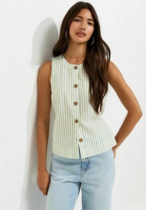 Femme aux longs cheveux foncés portant des boucles d'oreilles créoles, un haut sans manches à rayures verticales vertes et blanches boutonné, et un jean bleu clair, debout contre un mur blanc.