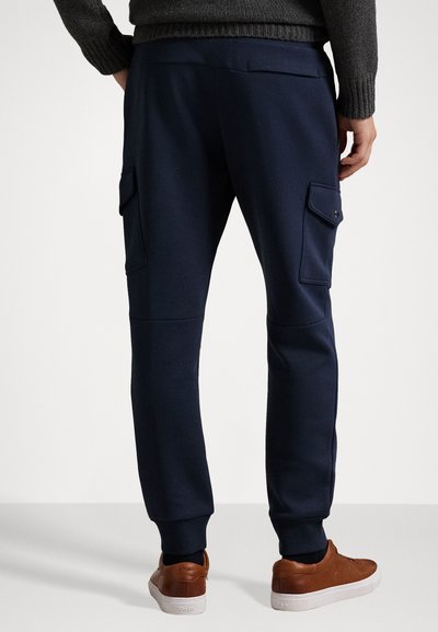 Bărbat îmbrăcat cu o pereche de pantaloni jogger cargo bleumarin, cu buzunare laterale, un pulover gri închis și teniși maroon, stând în fața unui fundal alb.