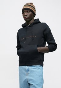 Jauns vīrietis valkā melnu "JAC & JONES" kapuci, gaiši zilas džinsus un brūnu svītrainu adītu galvassegu, ar rokām kapuces kabatā.