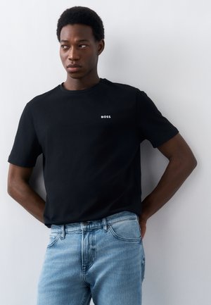 Άνδρας που φοράει μαύρο T-shirt BOSS και ανοιχτό μπλε τζιν, στέκεται μπροστά σε ένα λευκό τοίχο με τα χέρια πίσω από την πλάτη του.