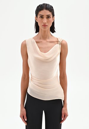 Donna con capelli effetto bagnato indossa un top crema senza maniche drappeggiato con spilla decorativa e pantaloni neri su sfondo bianco uniforme.