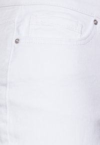 Primer plano de una tela de denim blanca que presenta un bolsillo trasero con una pequeña tachuela decorativa de color plata. Textura suave con detalles de costura.