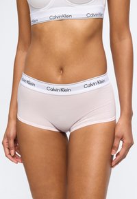 Γυναίκα που φοράει ανοιχτόροζ εσώρουχο boyshort Calvin Klein και ταιριαστό λευκό bralette Calvin Klein, και τα δύο με branded ελαστικές ζώνες στη μέση.
