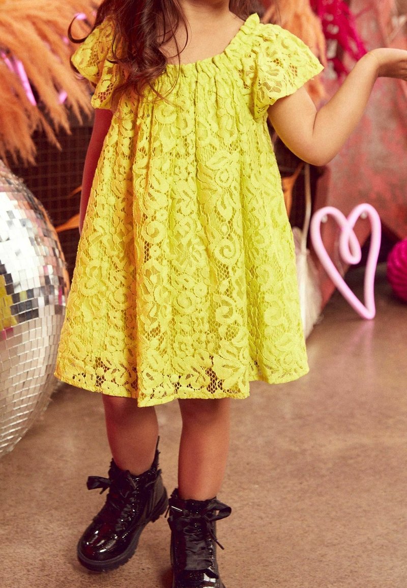 Enfant portant une robe en dentelle jaune et des bottes vernies noires debout à l'intérieur près d'une boule disco et d'une décoration en forme de cœur rose.