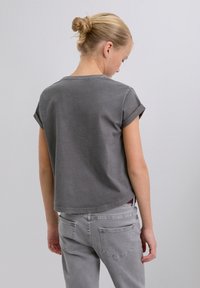 Grå bomulls-T-shirt med uppvikta korta ärmar, rundad halsringning och en boxig passform, sedd bakifrån tillsammans med ljusgrå jeans.