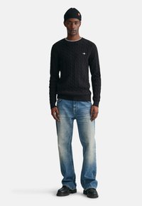 Schwarzer Strickpullover mit rundem Ausschnitt, kombiniert mit hellblauen geraden Jeans und schwarzen klobigen Schuhen. Schwarze Mütze mit Markenlogo.
