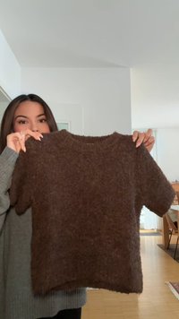 Donna con capelli scuri tiene in mano un maglione marrone a maniche corte e peloso, stando in una stanza luminosa con pavimenti e mobili in legno.