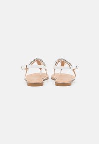 White sandals with a flat sole, featuring a beaded upper design and adjustable ankle strap. Smooth texture with a light brown insole.