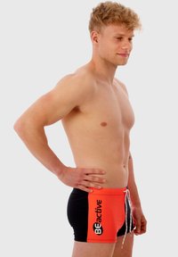 Shorts de bain rouges et noirs avec cordon de serrage, portant le texte "BEactive" sur le côté. Fabriqués en tissu lisse et extensible.