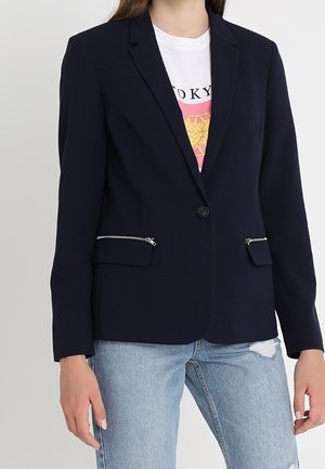 Persona con una chaqueta azul marino con bolsillos delanteros con cremallera, sobre una camiseta blanca con gráfico y vaqueros rotos azul claro.