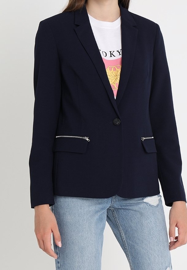 Personne portant un blazer bleu marine avec des poches avant zippées sur un t-shirt blanc à motif graphique et un jean bleu clair déchiré.