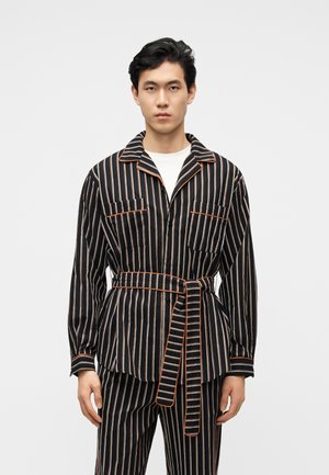 Άνδρας που φοράει μαύρο και μπεζ μπουφάν με κάθετες ρίγες και ζώνη, καθώς και παντελόνι που ταιριάζει, στέκεται μπροστά σε απλό ανοιχτό φόντο.