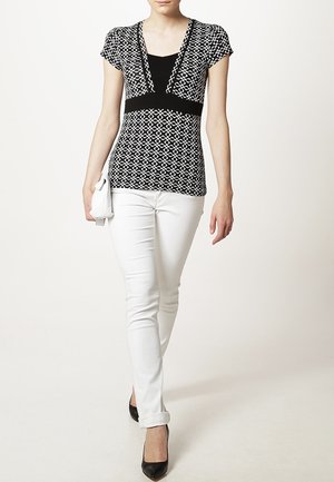 Czarnobiała wzorzysta bluzka z krótkimi rękawami, dekoltem w kształcie litery V i gładkim czarnym środkowym panelem, zestawiona z białymi dopasowanymi spodniami i czarnymi szpilkami.
