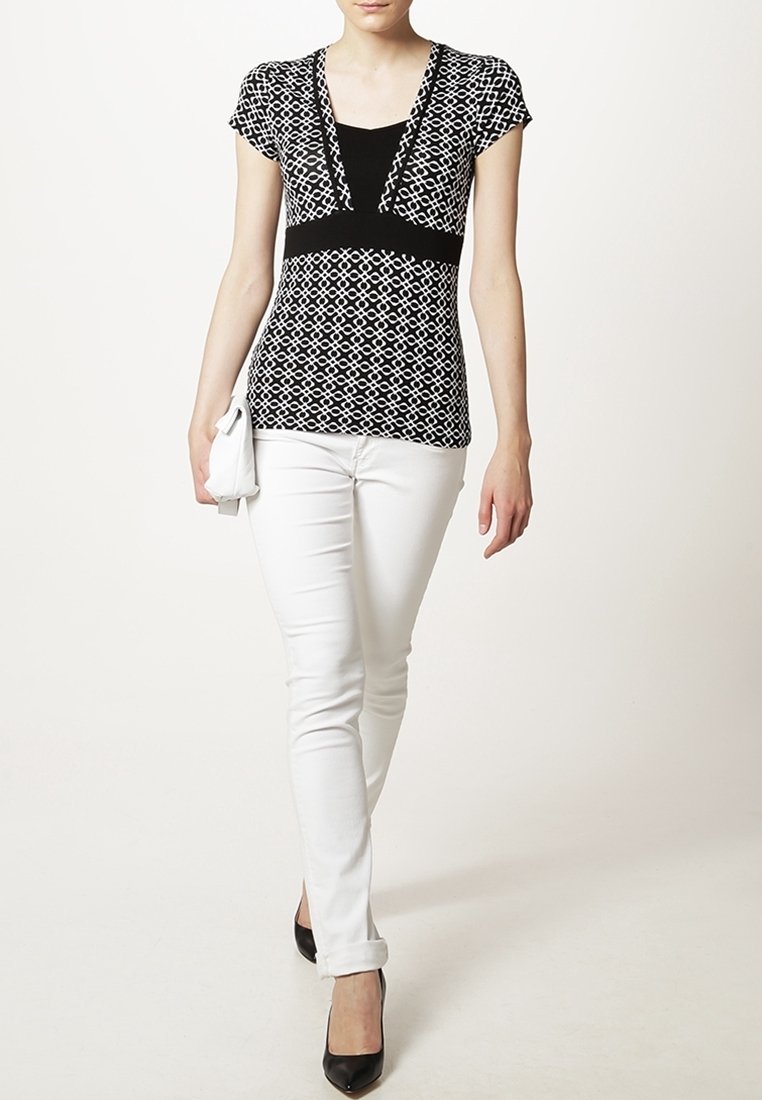 Haut à motifs noir et blanc avec manches courtes, décolleté en V, section centrale noire unie, associé à un pantalon blanc ajusté et des talons noirs.