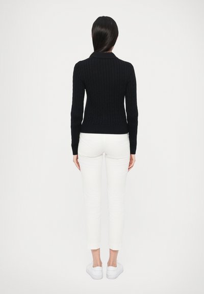 Tmavomodrý pletený sveter s rebrovanou textúrou, priliehavým strihom a dlhými rukávmi, v kombinácii s bielymi úzkymi nohavicami a bielymi teniskami.