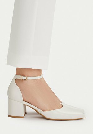 Fuß mit weißem Schuh mit Blockabsatz, Knöchelriemen und spitz zulaufender Spitze, kombiniert mit weißen kurzen Hosen.