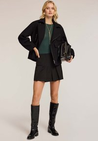 Zwarte cropped jas, groene top, zwarte geplooide rok en kniehoge glanzende zwarte laarzen. Accessoires zijn een zwarte tas en een gouden ketting.