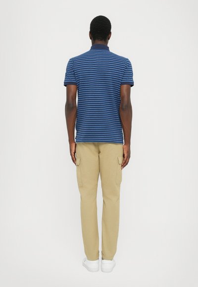 Tums zilas svītrains polo krekls apvienots ar khaki krāsas īsām biksēm. Nodrošina ikdienišķu piegriezumu ar īsām piedurknēm un apkakles detaļu. Redzamas baltas kedas.