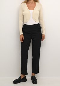 Hellbeige gekürzte Strickjacke mit langen Ärmeln und Stricktextur. Kombiniert mit hochtaillierten schwarzen Hosen und schwarzen Loafers. Einfaches Design.