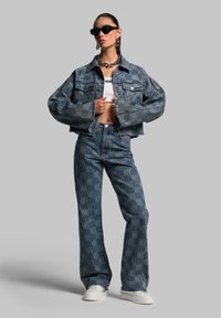 Denim jacket and matching jeans in faded blue featuring large pockets and a repeated logo pattern. Model wears sunglasses and white shoes.