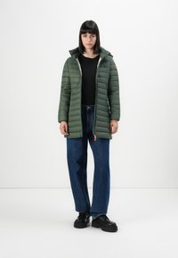 Vrouw in een groene gevoerde jas, zwarte shirt, blauwe jeans en zwarte robuuste schoenen, staand tegen een witte achtergrond.