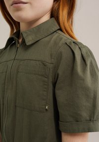 Camicia a maniche corte verde oliva con zip frontale, due tasche sul petto, dettagli plissettati sulle spalle e una piccola stella decorativa sulla manica.