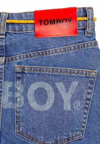 Denimjeans met een felrode label met "TOMBOY" in zwart, gele stiksels en een klassiek vijfzakkenontwerp.