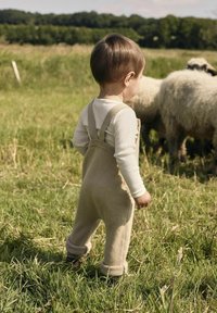 Beige gestrickte Latzhose mit verstellbaren Trägern, kombiniert mit einem weißen Langarmshirt. Das Kind steht im Gras mit Schafen im Hintergrund.