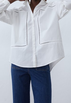 Persona con camisa blanca de manga larga con botones, con cortes verticales decorativos y vaqueros azules, ajustando el cuello.