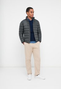 Giacca puffa grigia, maglione blu navy, camicia azzurro chiaro, pantaloni beige e sneakers bianchi. Texture liscia, chiusura a zip e motivo trapuntato orizzontale.