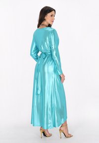 Teal satin lång klänning med lång ärm och midjeband, som har en mjuk textur och uttalad kjol, bärs med guldfärgade högklackade sandaler.