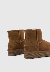 Botines de ante marrón con suelas de goma planas, costuras visibles y forro suave en su interior, mostrados desde la parte trasera sobre un fondo liso.