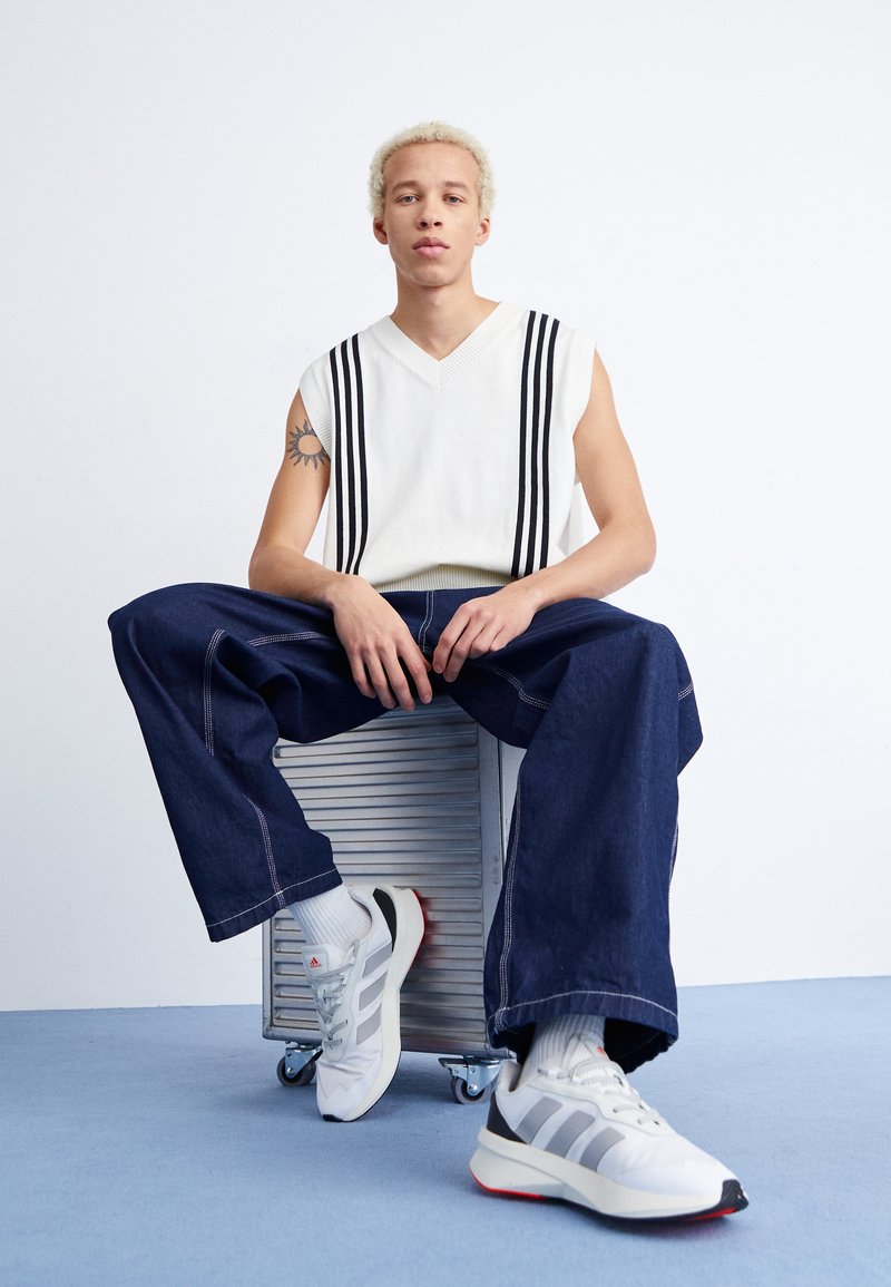 Maglione bianco senza maniche con strisce verticali nere, abbinato a jeans larghi in denim scuro e sneakers grigie chiare con dettagli rossi.