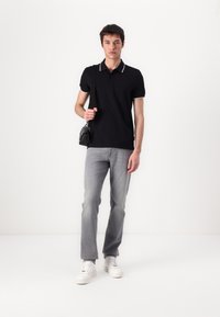 Čierne polo tričko s bielymi detailmi na golieri, šedé priame džínsy a biele tenisky. Model drží malú čiernu tašku. Minimalistický dizajn.