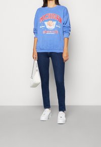 Sudadera azul claro con "MICHIGAN" y gráfico de baloncesto, combinada con jeans azul oscuro y zapatillas blancas. Sosteniendo un bolso blanco.