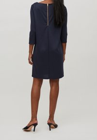 Robe bleu marine avec manches trois-quarts, un col arrondi et un détail de fermeture éclair dorée dans le dos. Texture lisse, coupe longueur genou.