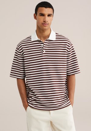 Ung mand med kort mørkt hår iført en brun og hvidstribet poloshirt og lyse bukser mod en ensfarvet baggrund.